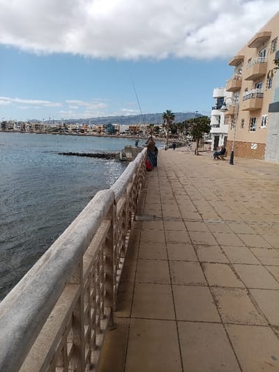 The image displays a daytime scene along the coastal promenade of Playa de Arinaga, located within the municipality of Agüimes, Gran Canaria, Spain. The perspective is longitudinal, with the ocean on the left and urban structures lining the right.

In the immediate foreground, a weathered, light-colored concrete railing with a square lattice pattern forms a barrier along the water's edge. The promenade surface consists of square, light brown paving tiles, exhibiting localized wear and discoloration.

The midground features the dark blue-green ocean with minimal wave activity. A short, rocky breakwater protrudes into the water from the shore. Two individuals are positioned at the railing, engaged in fishing; one person, wearing a dark jacket and pants, holds a fishing rod extending over the water, with a red bag at their feet. Another individual is partially obscured behind them. Further down the promenade, several indistinct figures are visible, some appearing to walk. Sparse palm trees and street lamps are present along the walkway. A solitary figure is seated on a bench to the far right.

The right side of the image is dominated by multi-story buildings, including a prominent light beige structure with multiple white balconies. These buildings extend into the background, forming a continuous coastal townscape. Distant hills or mountains are discernible beyond the urban area. The sky above is light blue with scattered white cumulus clouds.