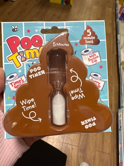A person's hand, wearing a pink ribbed long-sleeved shirt, holds a novelty "Poo Timer" hourglass toy. The toy is shaped like a brown stylized pile of feces and is encased in clear plastic packaging with a blue checkered background. Text on the packaging includes "Poo Timer," "5 Minute Limit," "With Suction Cup," and "Wipe Time!". The background suggests an indoor setting, possibly a retail environment, with wooden flooring visible. There is no discernible indication of the city of Carlow, Ireland, within the image.