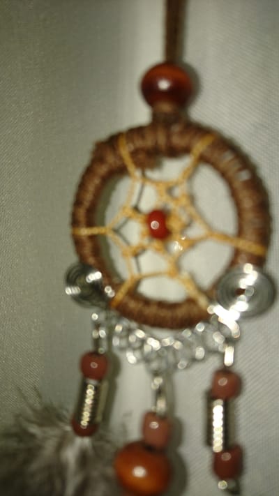 The image shows a close-up, out-of-focus view of a dreamcatcher. The dreamcatcher is made of a circular frame wrapped in brown string, with a yellow web pattern inside. A red bead is at the center of the web. Silver decorative elements and red beads hang from the bottom of the dreamcatcher. A small section of a gray feather is visible to the left. The background is a light-colored, textured surface, possibly fabric. There is no discernible information within the image to indicate a location in Dvirets, Ukraine.