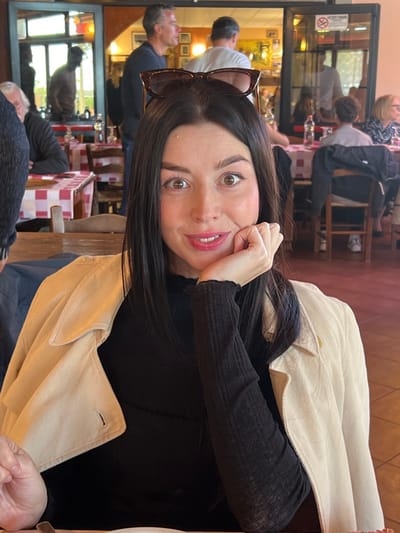 The image depicts a young adult female seated in a restaurant in Civitavecchia, Italy, captured in a waist-up, direct-gaze portrait. She has long, dark brown hair, centrally parted, with tortoiseshell-patterned sunglasses perched on top of her head. Her eyes are wide and brown, looking directly at the viewer, and her lips are slightly parted in a faint smile, revealing pink coloration. She is wearing a black, long-sleeved ribbed top, with a light beige/tan corduroy jacket draped over her shoulders. Her left hand is positioned with fingers gently curled under her chin.

The background reveals a bustling restaurant interior. Multiple wooden tables are present, covered with red and white checkered tablecloths. Traditional wooden chairs with rush seats are visible. Several other patrons are discernible in various states of activity: an older male with white hair is partially visible to the left, a man in a dark striped shirt stands looking towards the right, another man in a white t-shirt stands with his back partially to the camera, and a woman with lighter hair is seated at a table further back on the right. The walls are light-colored, with framed items visible in the distance. A large doorway or archway separates sections of the restaurant, with glass panels present on the right side of this opening. A red "NO SMOKING" sign is affixed above the doorway. Plastic water bottles and glasses are visible on some tables. The floor is tiled in a warm, brownish-orange hue. The lighting suggests a bright, possibly daytime, interior.