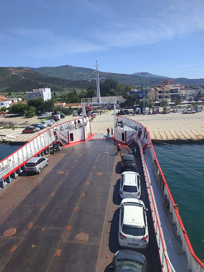 The image shows a ferry docked at the port of Prinos, Greece, under a clear blue sky. The ferry's deck is occupied by several cars, including a silver Fiat Punto, a black hatchback, and three white hatchbacks parked in a line. A motorcycle is also visible on the deck. A red and white railing runs along the side of the ferry. In the background, the town of Prinos is visible with buildings and lush green hills. Several people are on the ferry, some appearing to be crew members or passengers. The water is a deep blue, indicating the sea. The overall scene is one of a typical ferry operation in a coastal town.