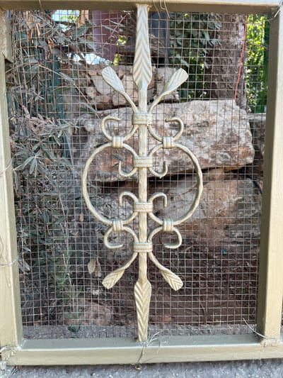 The image displays a close-up, vertical view of a light green or gold-painted metal gate or fence panel, located in the Menashe Regional Council, Israel. The panel features a central, ornate decorative element composed of scrolled and leaf-like motifs, flanked by a fine, silver-colored wire mesh with a square grid pattern. The metal frame of the panel is also painted the same light green/gold color, with minor paint drips visible, particularly at the bottom. Behind the mesh, the environment consists of large, light brown rock formations, sparse green foliage including shrubs with narrow leaves on the left, and a darker, possibly shaded area with denser vegetation on the right. The ground visible through the mesh appears to be dry soil or dirt. The scene is illuminated by natural daylight. No individuals or animals are present.