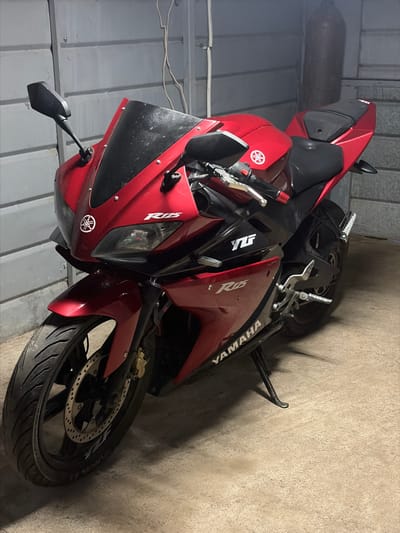 A red Yamaha R15 motorcycle is parked inside a building in Zimnitsa, Bulgaria. The bike is positioned diagonally, facing left, with the front wheel visible in the lower right corner. The left side mirror is extended. The building walls appear to be made of corrugated metal, while the floor is concrete.