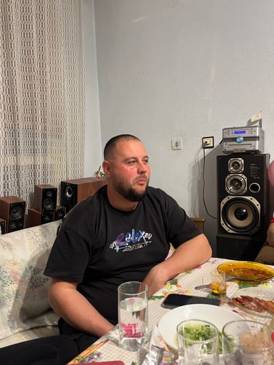 A man is seated at a table covered with a patterned tablecloth. There are various plates of food and drinking glasses on the table. He is wearing a black t-shirt with colorful graphics. Behind him, there is a wall with a window covered by a white curtain. There is a stereo system with speakers mounted on the wall. Further behind him is a stack of speakers. The scene appears to be indoors, likely in a dining area. It is not possible to determine the location in Pamukchii, Bulgaria, based on the information in the image.