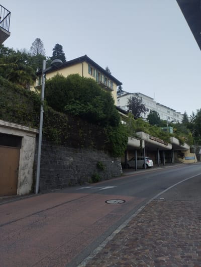The image depicts a view of a street in Orselina, Switzerland, with buildings and vegetation. In the foreground, a road with a red asphalt surface is visible, leading into a gray asphalt road. A cobblestone sidewalk lines the right side of the road. On the left, a stone wall covered in vegetation supports a building with a beige facade and green shutters. A metal railing is also visible on the upper left. A streetlamp stands tall next to the building.
Further back, a multi-story building with a white facade and many windows is visible, possibly a hotel or residential complex, set against a slightly hazy sky. Below this building, a series of covered parking spaces or terraces with curved concrete structures, also overgrown with plants, are present. A white car is parked in one of these spaces. The overall impression is that of a residential or commercial area on a hillside, with a focus on integrating architecture with nature.