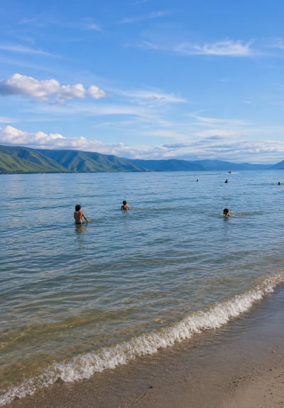 The image depicts a serene, natural landscape with a body of water and distant mountains. Several children are visible in the water, with some wading and others swimming. The water is clear, with gentle waves lapping at the sandy shore in the foreground. The sky is a bright blue, dotted with fluffy white clouds. Mountains with lush green slopes rise in the background, forming a picturesque backdrop. A small boat is visible in the distance on the water. The scene appears to be a tranquil day at a lake or sea, likely in a location like Situngkir, Indonesia, known for its natural beauty.