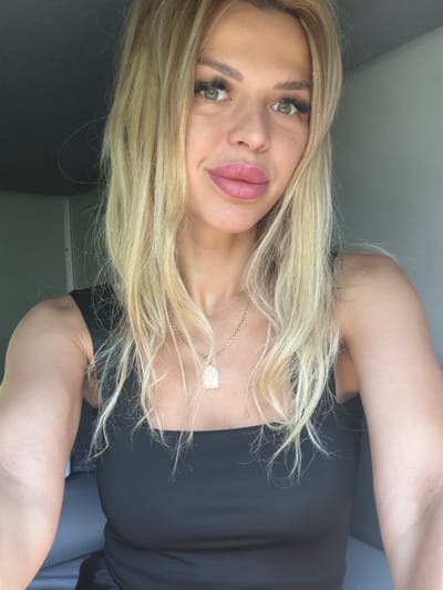 This is a close-up, rotated selfie of a blonde woman. She is wearing a black top and a necklace with a rectangular pendant. Her hair is long and blonde, and it falls over her face and shoulder. Her eyes are green, and she has full lips with pink lipstick. The background is a plain gray wall. The lighting suggests it is daytime. No text is visible in the image.