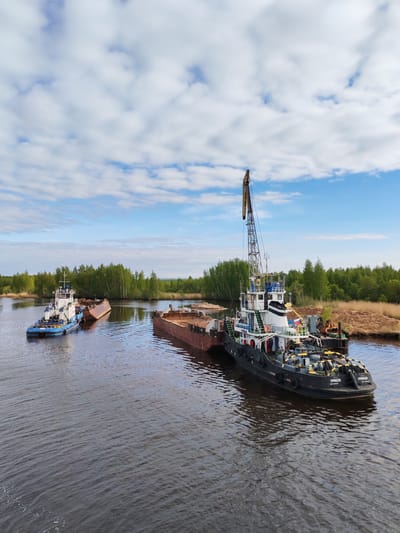 The scene depicts a waterway in Ozernoe Ust'e, Russia, with two tugboats and barges. The larger tugboat, primarily black with white accents, is equipped with a crane and is docked alongside a barge loaded with what appears to be sand or gravel. The tugboat has the inscription "ПРИБОЙ" on its hull. A smaller, blue and white tugboat is visible in the distance, towing another barge which seems partially submerged. The waterway is surrounded by green trees and vegetation on both banks, suggesting a rural or natural environment near an industrial operation. The sky is blue with scattered white clouds, indicating a fair weather day.