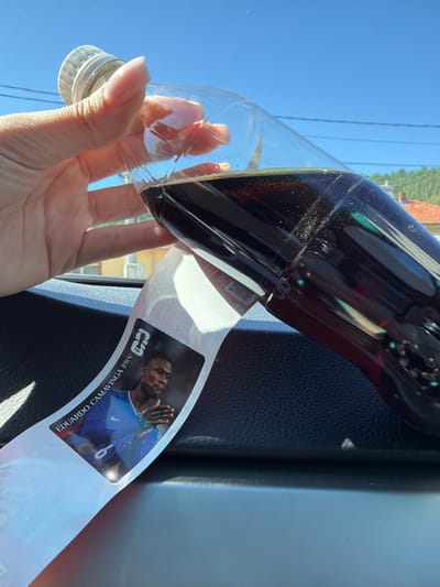 A hand holds a partially filled plastic bottle of dark soda, tilted forward, with the bottle's label featuring a picture of a soccer player, Eduardo Camavinga (FRA). The bottle is positioned over a black textured surface, likely a car's dashboard. In the background, a bright blue sky is visible with subtle power lines and distant greenery with red-tiled roofs, suggesting an outdoor, possibly urban or suburban setting. The image does not contain information indicating the specific location within the city of Beli Osam, Bulgaria.