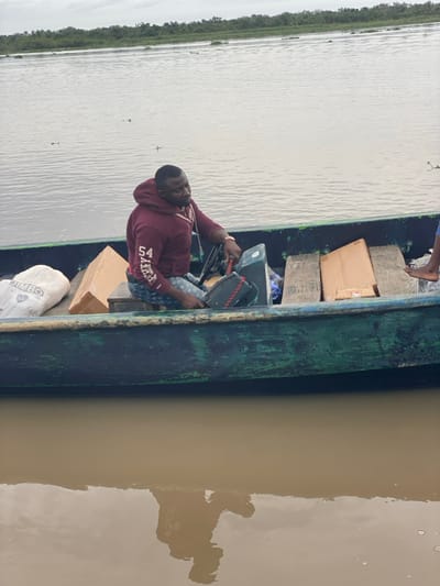 A dark green, weathered wooden longboat is afloat on muddy brown water in a waterway near Akarakumoh, Nigeria. The background features calmer greyish water and a dense line of green vegetation under an overcast sky. A dark-skinned adult male with a short haircut and beard is seated in the boat, facing right. He wears a maroon hooded sweatshirt with "54 JAMBO" printed on the left sleeve and patterned blue shorts, along with a gold-colored watch on his left wrist. His hands are positioned on the controls of a dark grey outboard motor unit with a red kill switch lanyard. Inside the boat, a white sack labeled "JIMBO" is visible on the left, and several brown cardboard boxes are stacked on the right. A bare foot, belonging to an unseen person, is partially visible at the far right of the boat. The reflection of the man and boat is visible in the brown water.