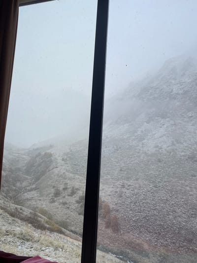 The image displays a snow-dusted mountainous landscape viewed from indoors through a window. A dark, thick vertical window mullion divides the scene centrally. To the left, a partial brown curtain frames the interior edge. A small area of reddish-pink fabric is visible along the bottom edge of the frame.

The exterior scene reveals a valley on the left, featuring descending slopes covered in sparse, snow-dusted vegetation, including pale grasses and scattered small trees with reddish-brown foliage. A narrow stream or river winds through the valley floor at the bottom. On the right, a steep, rocky mountain face rises, also covered with a light dusting of snow. Patches of fog or low-hanging clouds obscure sections of the mountain face and the upper portions of the valley, creating a misty atmosphere. The sky is uniformly overcast and light gray. Numerous small white specks are visible across the entire outdoor view, indicating light snowfall. No individuals, animals, or specific structures are discernible within the outdoor environment. This scene is consistent with the high-altitude, rugged terrain found near Juta, Georgia, during winter or late autumn.
