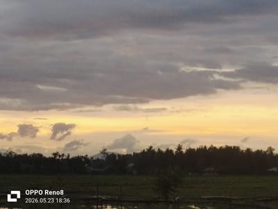 The image depicts a twilight scene in Kampungjumpa, Indonesia. The foreground shows a grassy field. A dark treeline extends across the midground, with silhouettes of various trees, including palm trees, against a vibrant, multicolored sky. The sky transitions from gray and purple at the top to orange and yellow near the horizon. Cloud formations are visible throughout. In the bottom left corner, there is a watermark indicating the image was taken with an OPPO Reno8 on May 23, 2026, at 18:36.