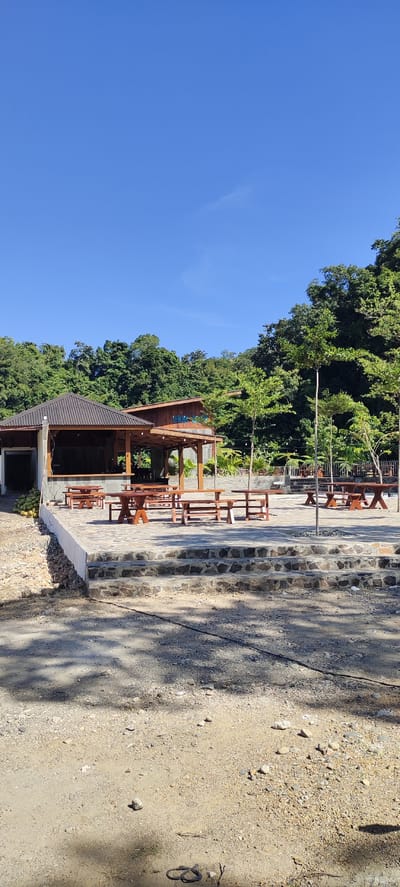 The image depicts an outdoor seating area in front of a wooden structure, likely a restaurant or cafe, located in Lamseunia, Indonesia. The area features multiple picnic tables and benches made of wood, arranged on a paved patio area. The patio is accessed via stone steps. Behind the seating area, a dense, green forest covers a hillside under a clear blue sky. The scene is bathed in sunlight, casting shadows on the ground. There are no people visible in the image. The overall impression is of a relaxed, open-air dining or gathering space surrounded by natural beauty.