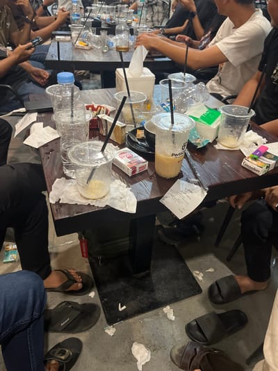 The image depicts a cluttered table in what appears to be a cafe or similar establishment in Meunasahceubrek, Indonesia, as evidenced by the "Pertamina Coffee" receipt. The table is covered with numerous empty plastic cups, some with straws, and empty water bottles. There are also cigarette packs on the table, including brands like "Commodore" and "Lucky Strike". Pieces of crumpled tissue paper are scattered around. People are seated around the table, partially visible in the image. Some individuals are using their phones. The floor around the table is concrete and has discarded tissue and debris. There are various types of footwear visible, including flip-flops and sandals.