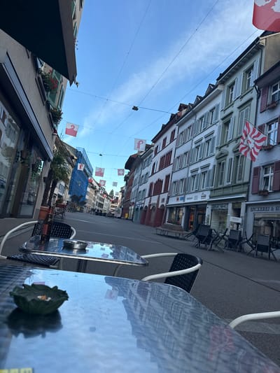 The image depicts a vibrant street scene in Liestal, Switzerland, characterized by traditional European architecture and outdoor cafe seating. The perspective is from a cafe table, looking down a street lined with multi-story buildings on both sides.
No people are visible, but the setting suggests a potential for outdoor dining or leisure. Numerous flags adorn the street, strung across overhead wires, prominently featuring Swiss flags (white cross on red) and flags of the Canton of Basel-Landschaft (red and white stripes with a black staff). One flag also shows a red and white striped pattern with a chicken-like emblem, likely representing Liestal itself.
The buildings feature various architectural details, including multiple windows with traditional shutters, some red and some green. Shopfronts are visible at street level. On the right, a building displays signage with "BÄCKEREI & KO..." (likely "Bäckerei & Konditorei" - Bakery & Confectionery) and "Malergeschäft" (Painting business). Another building on the left has signs for "FUTURO" and "Innovatis". Further down the street, scaffolding covered with blue netting is visible on a building, indicating ongoing construction or renovation.
In the foreground, two outdoor cafe tables are present, one with a reflective surface showing reflections of the buildings and flags. An ashtray sits on one table, and a crumpled dark green leaf rests on the other. Black chairs with light-colored frames accompany the tables. The street surface is paved with asphalt.
The time of day is clearly daytime, under a bright, clear blue sky with minimal cloud cover, suggesting pleasant weather. The overall impression is one of a calm, well-maintained town street, possibly captured during a quiet moment.