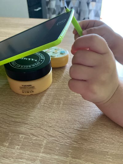 A close-up shot shows a child's hands interacting with a digital drawing tablet and a jar of cream. The tablet, with a green frame, is tilted and positioned on top of a jar labeled "YASMIN" and "КРЕМ-ВОСК ОТ ТРЕЩИН" (Cream-wax for cracks). The jar itself is yellow and has a black lid. A small, yellow toy with buttons is also visible near the tablet. The child's hands are holding the green stylus of the tablet and are close to the drawing surface. The scene is set on a wooden table. The image appears to be taken indoors, possibly in a home environment, in the city of Maksimkovo, Russia.