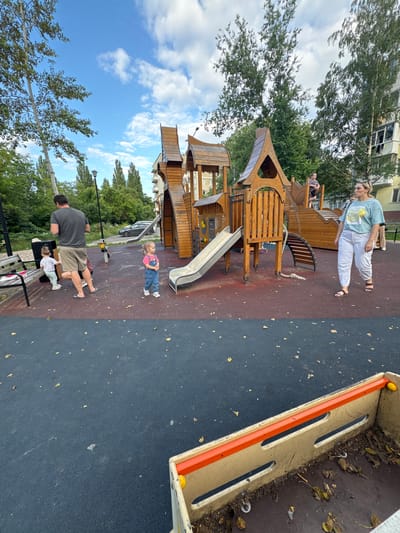 The image captures a scene at a children's playground in Vyazniki, Russia. The central focus is a large wooden playground structure with multiple levels, a slide, and wooden stairs. In the foreground, a man and two young children are present. The man, wearing a grey t-shirt and khaki shorts, is standing near a park bench. One child, wearing a white shirt and light-colored pants, is sitting on the bench. Another child, in a pink shirt and blue overalls, is standing near the playground structure.

Further back, a woman in a light blue t-shirt and white pants is standing. She appears to be looking down. Another child is visible on a higher platform of the playground structure, wearing a dark and white patterned shirt and dark shorts.

The playground is situated on a dark, rubberized surface, with scattered dry leaves. Beyond the playground, trees and a residential building are visible. The sky is blue with scattered white clouds, suggesting it is daytime. The overall atmosphere appears to be casual, indicative of a typical day at a park. There are no visible text elements in the image.