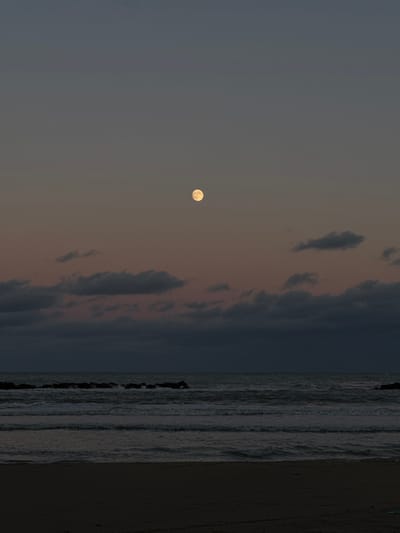 This image depicts a coastal scene in Grottammare, Italy, at twilight. The sky transitions from a deep charcoal grey at the top to a muted reddish-grey above the horizon. A prominent, pale yellow full moon is positioned in the upper center of the frame. Below the moon, fragmented, dark grey clouds are distributed across the mid-sky. The sea exhibits a dark, agitated surface with discernible wave formations and intermittent whitecaps. A horizontal line of dark, irregular rock breakwaters is visible approximately one-third of the way up from the bottom of the image, interacting with incoming waves. The immediate foreground consists of a dark, indistinct sandy beach. No human figures or discernible man-made objects are present on the beach or in the water.