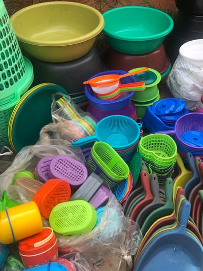 In Amaeke Abam, Nigeria, an eye-level shot reveals a jumbled pile of colorful, plastic household items outdoors, including bowls, dustpans, strainers, and buckets in varying sizes and shapes. The items are of various colors, including green, yellow, orange, red, blue, and purple. A white plastic bag is also visible in the pile. The scene is set on the ground, possibly dirt or grass.