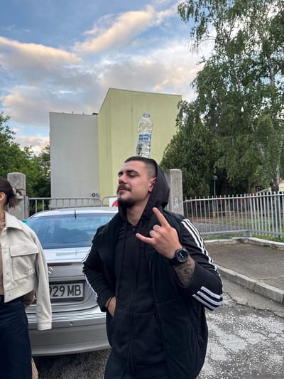 A man balances a water bottle on his head while standing in front of a silver Mercedes-Benz in Пловдив, Bulgaria. The man is wearing a black Adidas tracksuit and a watch on his left wrist, which is adorned with a tattoo. He is making a "rock on" hand gesture. A woman wearing a white jacket and jeans is standing in front of the car. The background shows a building, trees, a fence, and a cloudy sky.
