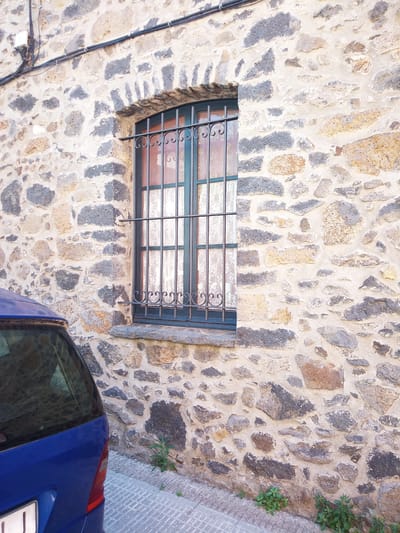 The image displays a section of a weathered stone wall, typical of the architecture in Castellfollit de la Roca, Spain. A small window, reinforced with a black metal grate featuring decorative scrollwork, is set into the wall. The window is fitted with lace curtains. To the left of the wall, the rear of a blue car is visible, parked on a patterned tiled walkway. Green plants grow along the base of the wall and the edge of the walkway. Above the window, electrical conduits and a junction box are attached to the wall. The scene is captured in daylight, with natural lighting illuminating the textured stone and surrounding elements.