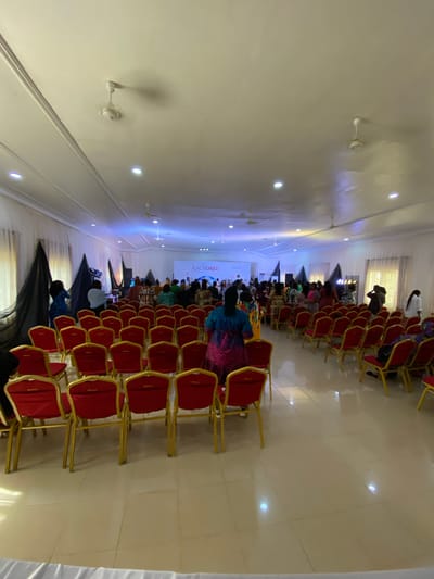 The image shows an event taking place in Miango, Nigeria. An audience sits in rows of red chairs facing a stage with a screen displaying the word "ANCHORED." Some audience members are visible wearing colorful clothing. Black drapes hang in the foreground and background. The room has a white ceiling with recessed lighting and ceiling fans. The floor appears to be tiled.