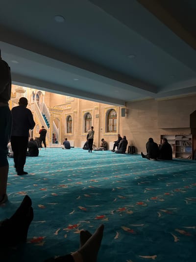 The image depicts the interior of a mosque in Breznitsa, Bulgaria. The foreground is dominated by a blue-green patterned carpet with orange, white, and brown floral or leaf motifs, observed from a low perspective showing the feet of an individual wearing black socks. Approximately 10-15 adult males are present within the large, carpeted space. Most individuals are seated on the carpet, some leaning against the light-colored, subtly textured walls on the right side. Clothing is generally casual and dark-toned, including sweaters, jackets, and trousers.
To the left, one man stands with his back to the viewer, while another in a light-colored hoodie walks barefoot across the carpet towards the left. On the far right, a simple white shelving unit holds books and miscellaneous items.
The background reveals an elaborately decorated inner courtyard or prayer hall section. This area features light brown walls with intricate gold and possibly blue decorative patterns, including arched windows with wooden frames. A prominent white and gold staircase with green-tiled steps ascends towards the left, leading to an upper level. The ceiling overhead is flat, light-colored, and contains recessed circular lights. The overall atmosphere appears calm, with individuals engaged in quiet contemplation or conversation.