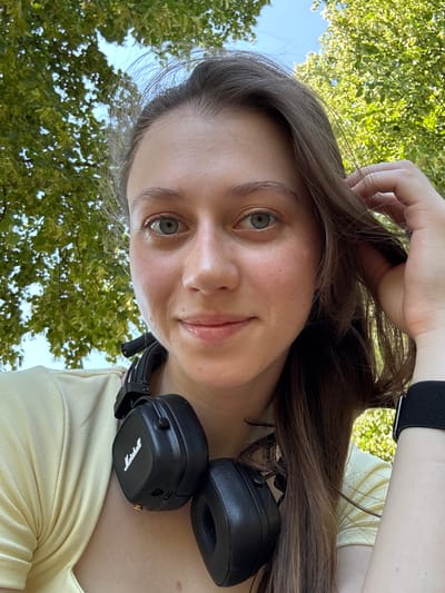This is a medium close-up, selfie-style portrait of a young woman with long, brown hair. She is outdoors, with green trees and a bright blue sky visible in the background. Her head is tilted slightly, and her left hand is touching her hair, resting on the top of her head. She is wearing a light yellow, ribbed top and a black smartwatch on her left wrist. A pair of black Marshall headphones is resting around her neck.
The woman has fair skin with some light blush on her cheeks and a subtle sheen on her lips. Her eyes are open and looking towards the camera, with a neutral or slightly curious expression. The lighting suggests it is daytime, likely sunny due to the bright sky and the illumination on her face and the foliage. The presence of leaves and trees implies a natural, outdoor setting, possibly a park or a garden, given the urban context of Erlenbach am Main, Germany. No specific activity is evident beyond posing for a photo. The visible text on the headphones reads "Marshall".