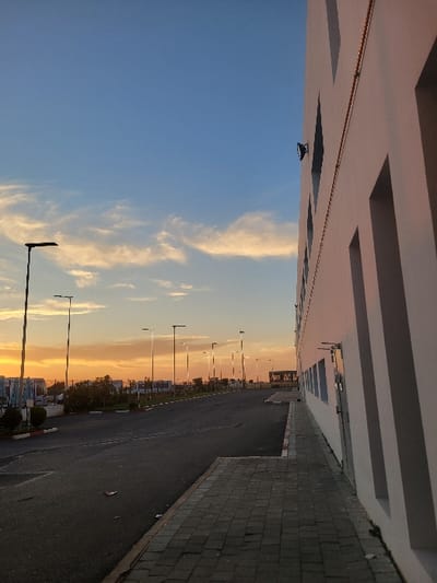A vertical view of a street during sunset. A white building with horizontal stripes dominates the top. A blue and orange sky is filled with clouds. The street is dark and contrasts with the sky, bordered by a gray brick walkway. Streetlights line the road, fading into the distance. Distant buildings are visible on the right-hand side of the image.