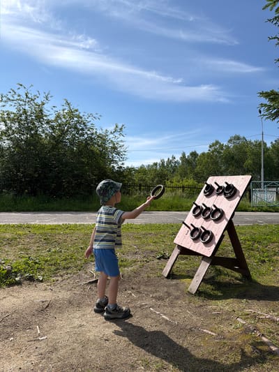 A young boy, facing away from the camera and towards a ring toss game, stands in a grassy, outdoor area under a bright blue sky with wispy clouds. He is wearing a striped t-shirt, blue shorts, a patterned cap, and hiking shoes. He holds a dark ring in his right hand, poised as if to throw it at the game. The ring toss game is a wooden board, painted pink, propped up on two A-frame wooden legs. It has nine black pegs arranged in three rows, designed to catch the thrown rings. The background features a dense line of green trees, a paved path, and a metal fence. The ground is a mixture of dirt and grass, with some scattered twigs. A lamppost is visible on the right side of the image. The scene is likely located in an outdoor recreational area or park in Kul'dur, Russia.