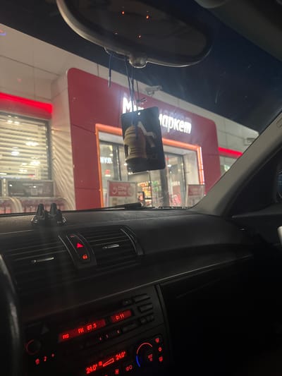 The image is taken from inside a car, looking out through the windshield at what appears to be a "Маркет" (Market) store. The store has a red and white exterior with glass windows. In the foreground, the interior of the car is visible, including the rearview mirror, dashboard air vents, and the car's radio/climate control panel. The radio display shows "FM 87.5" and a temperature of "24.0°". A card is hanging from the rearview mirror. The environment appears to be an urban or commercial area. The image does not include specific details to confirm the location as Vodenichane, Bulgaria. There are no people visible in the image.