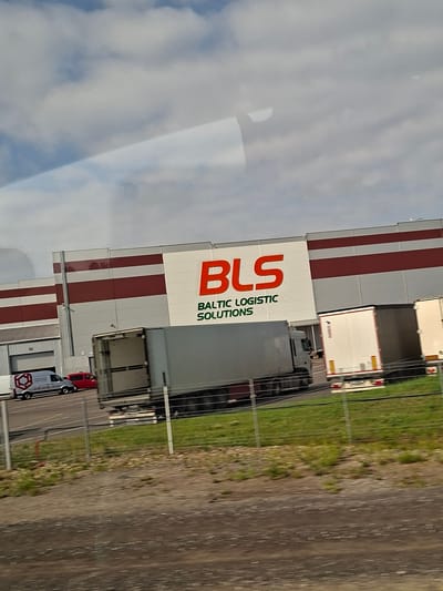 A medium shot captures the exterior of a modern warehouse facility with the prominent logo "BLS BALTIC LOGISTIC SOLUTIONS" displayed on its white facade. The building features horizontal bands of dark red and white. In the foreground, a chain-link fence runs across the bottom of the frame, with a strip of green grass and weeds behind it. To the left of the warehouse, a white delivery van with a hexagonal logo and the text "SERVISAS" is parked. Next to the van is a red passenger car. Several large trucks, including a semi-trailer truck with an open trailer, are parked in front of the warehouse loading bays. The sky above is a mix of white clouds and blue. The scene is a street-level view in Kampiškiai, Lithuania, suggesting a logistics or transportation hub.