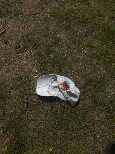 A white baseball cap lies on the ground amidst dry grass and dirt. The cap is creased and somewhat flattened, with its interior fabric visible. A patch on the side of the cap displays the German flag (black, red, and gold) and the word "DEUTSCHLAND". The surrounding ground is a mixture of sparse, dry grass, bare earth, and some twigs. There are no people or other objects of significance visible in the image. The lighting suggests it is daytime, with shadows indicating sunlight.