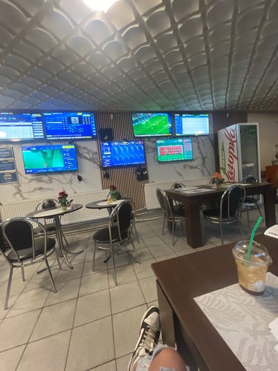 The image shows the interior of a sports bar or betting shop, likely located in Batoshevo, Bulgaria. The walls feature textured decorative panels, and numerous television screens display live sports events, possibly soccer, and betting information. Several small tables with metal chairs are arranged in the room, suggesting a casual seating layout. A plastic cup with a straw and some paperwork is visible on one of the tables, alongside part of a person's leg with sneakers. There is a refrigerator behind one of the tables.