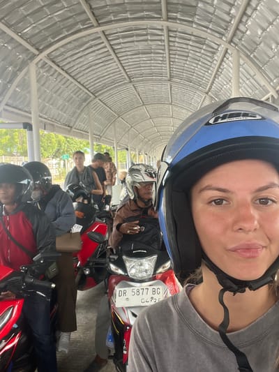 In the city of Upt Tambak Sari, Indonesia, a group of people are lined up on motorcycles under a covered walkway. A young woman in the foreground, wearing a blue helmet, is taking a selfie. The walkway has a corrugated metal roof supported by white posts. In the background, other individuals on motorcycles are visible, some wearing helmets. The scene appears to be an outdoor waiting area, possibly a transportation hub.