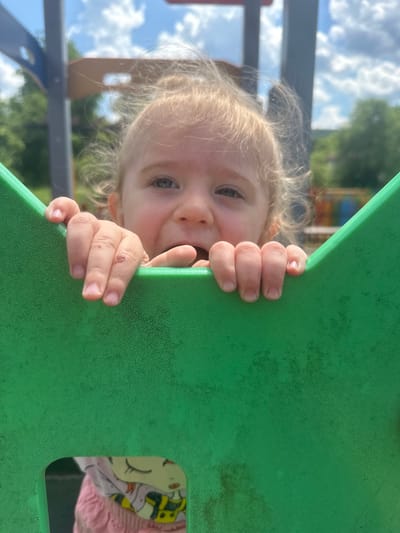 In Donchovci, Bulgaria, a close-up shot captures a young child peeking over a bright green plastic structure, possibly part of a playground. The child, with light-colored, wispy hair, looks directly at the camera with a wide-eyed expression. Their small hands, with visible fingernails and minor abrasions on one, grip the edge of the green structure. The background is blurred, showing a bright sky with scattered clouds and hints of green trees and playground equipment, suggesting an outdoor setting on a sunny day. A glimpse of another child's clothing, featuring a cartoon character and pink fabric, is visible through an opening in the green structure.