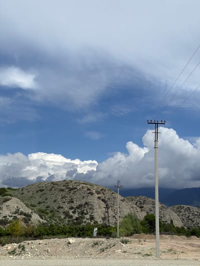 The image depicts a scenic view of Tamga, Kyrgyzstan, featuring rolling hills covered in sparse vegetation and gravel. A clear blue sky with scattered white clouds dominates the upper half of the frame. In the foreground, a concrete power pole with wires stretches diagonally across the image. Another, smaller power pole is visible in the mid-ground. The hills are characterized by their rocky terrain and patches of green shrubs. The overall scene conveys a sense of rural tranquility and natural beauty.