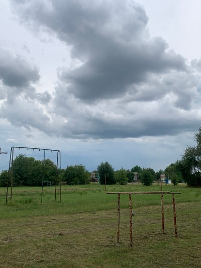 This image depicts an outdoor scene with a cloudy sky, featuring a grassy field with two sets of rusty metal exercise equipment. In the foreground, a pair of parallel bars stands on four legs. Further back, a horizontal bar with multiple hanging points is supported by two uprights. Trees and some small buildings are visible in the background. The scene appears to be located in a rural or semi-rural area, potentially a public space or an abandoned sports ground. The weather appears overcast, suggesting a potentially damp or cool day.