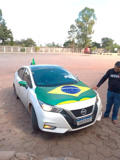 A white Nissan sedan is parked on a patterned paved surface in front of a concrete wall and trees. The car is decorated with a large Brazilian flag design covering its hood and a smaller Brazilian flag on its side mirror. A small triangular Brazilian flag is also attached to the roof of the car. A man wearing a black long-sleeved shirt and blue jeans is standing to the right of the car, looking down at the hood and resting his hand on it. The license plate of the car reads "SG05D92" and has "BRASIL" and "BR" printed on it. In the background, a red semi-trailer truck is visible. The scene appears to be an outdoor area, possibly a parking lot, with trees and a wall in the background.
