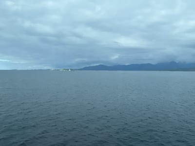 A wide shot shows a calm, dark blue sea stretching to the horizon under a cloudy, overcast sky. In the distance, along the horizon, a faint line of dark green land with subtle mountain outlines is visible, suggesting a coastal or island landscape. On the left side of the horizon, a small, white structure, possibly a lighthouse or building, stands out against the darker landmass. The water's surface is gently rippled, reflecting the muted light of the sky. The scene evokes a sense of quietude and vastness, typical of coastal Norwegian scenery.