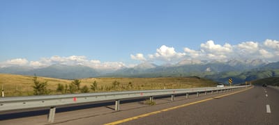 A roadside view captures a highway with a silver guardrail bordering the right lane. A white sedan is parked on the left side of the highway, and a black SUV is visible further down the road. The highway stretches towards a backdrop of rolling green hills and majestic mountains. The sky above is a clear blue, dotted with scattered white clouds. The scene is set in the vicinity of Қырғауылды, Kazakhstan, characterized by its natural mountainous landscape.