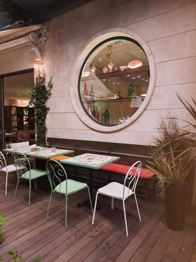 An outdoor seating area of a cafe or restaurant is depicted, likely in Pesaro, Italy, given the architectural style. The foreground features a wooden deck with several tables and chairs arranged for dining. The tables have decorative tops with floral patterns and are set with napkins and small glass containers. The chairs are a mix of light green metal with intricate scrollwork and white metal with simple, modern designs. Behind the tables, a long red bench seat is visible, suggesting additional seating.

The backdrop is a cream-colored, horizontally paneled wall. Dominating the wall is a large, circular window with a thick, light-colored frame. Inside this window, a reflection shows shelves lined with decorative glass bottles and other items, illuminated by warm lighting. A lush green vine climbs the wall near the left side of the image, adding a touch of nature to the scene. To the right of the circular window, a tall, dark planter holds a spiky, dry plant.

The overall ambiance suggests an evening setting, possibly after closing or during a quiet period, as no patrons are visible. The lighting is soft and atmospheric, enhancing the refined yet casual feel of the outdoor space.
