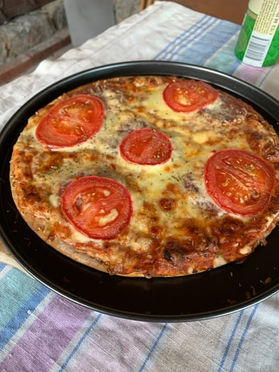 The image presents a freshly baked, round pizza served in a black, circular baking pan. The pizza features a golden-brown crust, a layer of melted white cheese that appears bubbly and slightly browned in areas, five distinct slices of red tomato evenly distributed, and a scattering of dried green herbs. Beneath the melted cheese, a reddish sauce base is visible, along with faint outlines of what appear to be small, round meat slices. The baking pan is positioned on a fabric tablecloth patterned with horizontal stripes in white, light blue, purple, and pink. In the blurred background, to the upper left, textured elements resembling a stone or brick wall and a white patterned fabric are visible. To the upper right, a portion of a tall, green beverage can or bottle is present, featuring a white label with a barcode and indistinct text, including the characters "CEN. AX. 5%". No individuals are depicted in the scene. The visual information within the image does not contain any details that would allow for the identification of a specific geographic location, such as Slokoshtitsa, Bulgaria.