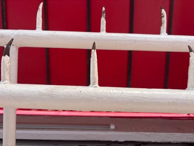 A close-up, low-angle shot captures a white, horizontally oriented metal railing with spiked protrusions, set against a bright red background. The railing appears to be part of a fence or barrier. The spikes are unevenly spaced and have a sharp, pointed appearance, some showing signs of wear or damage, revealing dark material beneath the white coating. The red background suggests a wall or structure, possibly outdoors. The scene is static and lacks any human presence. The lighting is even, with no strong shadows, indicating an overcast day or a shaded area. The overall impression is one of a protective or deterrent structure. The location is identified as Victoria, Morocco.