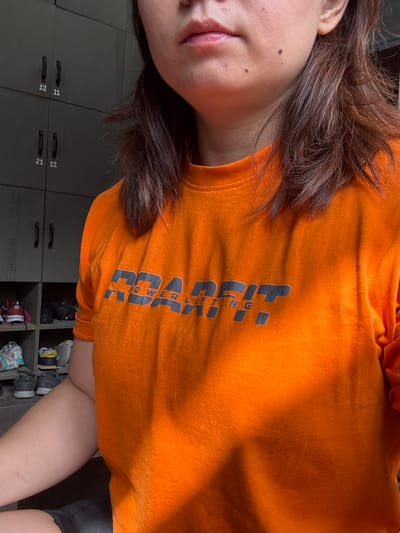 This media file is a close-up, low-angle shot of a person wearing an orange t-shirt with grey text. The subject's face is partially visible on the left side, with their mouth slightly open and hair falling across their face. The t-shirt has a bold, block-style graphic design that appears to be text, with the letters "POWERLIFTING" partially visible. The background shows a dark grey locker room with numbered compartments and shelves filled with shoes. The lighting suggests an indoor setting, possibly a gym or changing room. There are no clear indicators of time of day or weather. The overall impression is casual and personal. The text visible on the t-shirt reads "POWERLIFTING" in a stylized font. The lockers are labeled with numbers like "11," "12," "21," and "22."