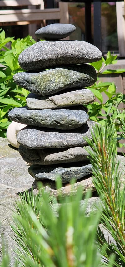 A stack of carefully balanced stones is positioned outdoors. The stones vary in size and shape, with a predominantly grey and greenish hue, some showing signs of moss or algae. The stack is in the foreground of the image, with lush green foliage in the background. Partially visible in the lower right corner are the needles of a pine tree, also in the foreground, slightly out of focus. Behind the foliage, a wooden structure, possibly a fence or furniture, is visible, with sunlight casting shadows. The overall setting appears to be a garden or natural outdoor space. The image does not contain any people or depict any actions or interactions. It is a still life composition focusing on the natural elements of stone and greenery.