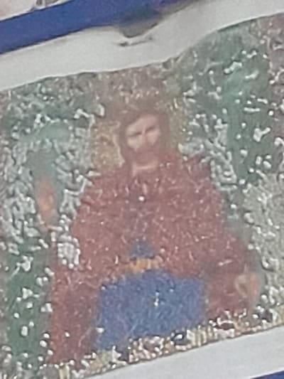 The image depicts a close-up, blurry view of a mosaic artwork. The central subject is a stylized depiction of a person, possibly religious in nature, with facial features and a beard visible. This figure is adorned in red and blue garments. The background is a textured green, resembling foliage or an abstract pattern. The mosaic appears to be framed by a white border, with a blue band visible at the top. The image quality is low, with significant pixelation and a lack of sharp detail. The location of this artwork within the city of Shostka, Ukraine, is not discernible from the visual information provided. There are no discernible actions or interactions occurring within the scene.