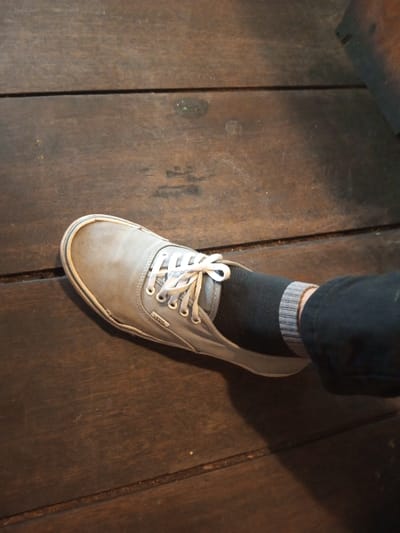 A grey Vans shoe sits on a dark wooden surface. The shoe is laced and shows a black sock with a grey striped cuff. A portion of a dark-colored pant leg is also visible. The lighting is slightly dim, highlighting the texture of the wood and the wear on the shoe. A portion of a wooden chair is visible in the upper-right corner.