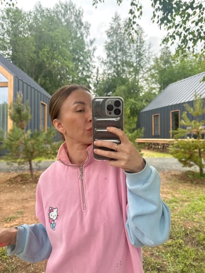 An adult woman with light brown hair pulled back takes a selfie with her smartphone. She is wearing a pink sweatshirt with light blue sleeves, featuring a small graphic of Hello Kitty holding a heart. The woman is standing outdoors in what appears to be a residential area with modern, dark-colored houses and scattered trees and greenery. The setting is identified as Novaya Vozhoika, Russia. She is holding the phone up to take the photo, with her lips pursed as if blowing a kiss. A ring is visible on her left hand. The background shows two houses, one on the left with a large window and a slanted roof, and another on the right with a darker, sloped roof. The ground is covered in dirt and grass, with some small pine trees and foliage. The sky is overcast.