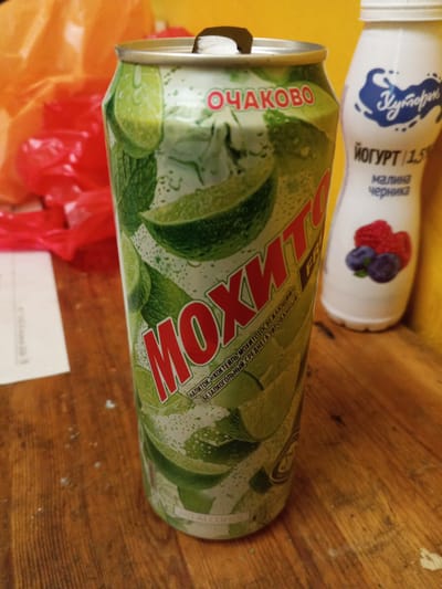 An eye-level medium shot shows a can of mojito-flavored beverage next to a bottle of yogurt. The mojito can has green and white packaging with images of limes and mint leaves. The text on the can is in Russian and appears to read "МОХИТО". The yogurt bottle is white with blue text and graphics, and it has images of raspberries and blueberries. The background is blurry and consists of orange and yellow objects, possibly plastic bags and a yellow wall. The scene is set on a wooden surface. The items are not arranged in any particular order, and there is no indication of their purpose or context. The lighting is dim and appears to be artificial. There is no visible sign of human presence or activity. The image lacks any geographical markers that would indicate its location within the city of Ulyanovka, Russia.